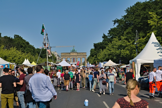 Eine Menschenmenge geht unter einem klaren blauen Himmel durch eine von Zelten, Fahrzeugen und B├Ąumen gesäumte Straße auf einen Bogen zu, mit Fahnenmasten auf der linken Seite, die auf das Oktoberfest in München, Deutschland, hinweisen.