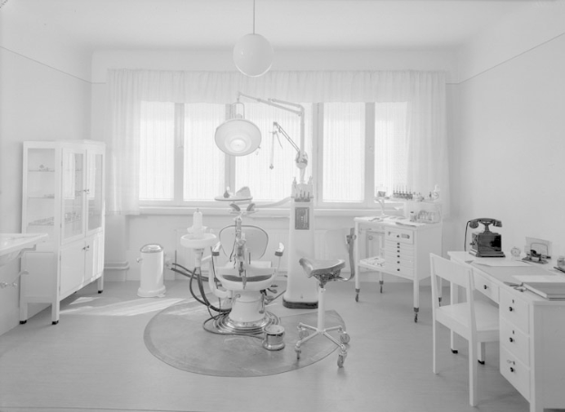 Ein Schwarz-Weiß-Foto einer Zahnarztpraxis mit einem Stuhl, einem Tisch, einem Schrank, einem Waschbecken, einem Mülleimer, einem Fenster mit Vorhängen und Deckenlampen, mit verschiedenen Gegenständen, die eine gemütliche und einladende Atmosphäre schaffen.