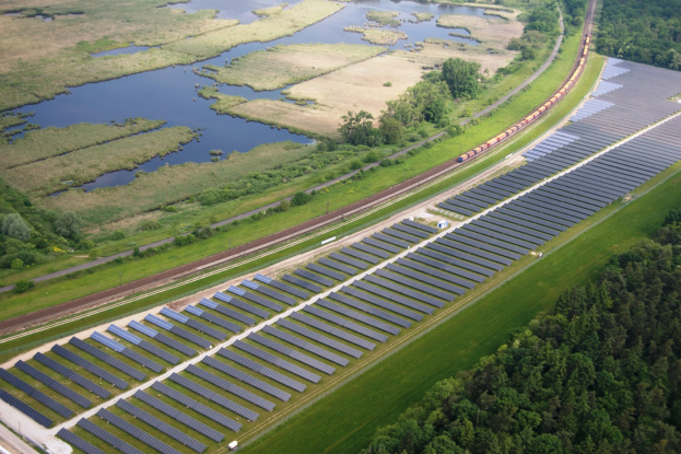 Luftaufnahme einer Solaränlage mit Panelen in einem Feld, umgeben von Bäumen, Gras, Pflanzen und Wasser, mit einem Zug auf einer näheren Eisenbahnschiene.