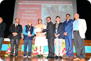 Eine Gruppe von Männern auf einer Bühne mit einem Zertifikat und einem Bildschirm im Hintergrund, der "Indian Institute of Food Processing Technology Awards 2019" und Blumendekoration zeigt.