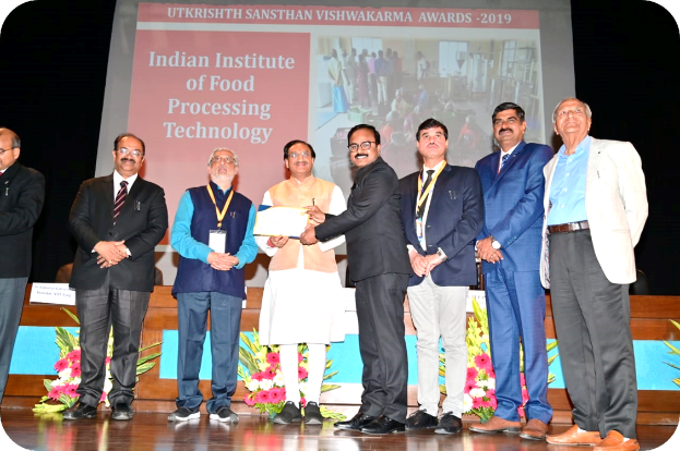 Eine Gruppe von Männern auf einer Bühne mit einem Zertifikat und einem Bildschirm im Hintergrund, der "Indian Institute of Food Processing Technology Awards 2019" und Blumendekoration zeigt.