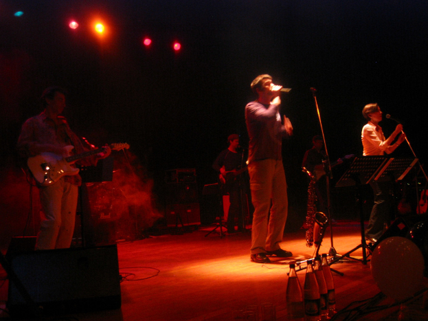 Zwei Menschen singen in ein Mikrofon auf einer Bühne mit drei Männern, die Gitarren spielen und Wasserflaschen auf dem Podium.