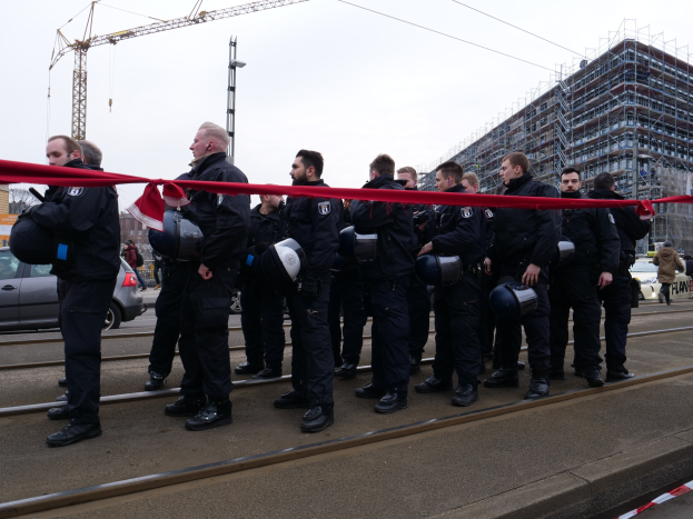 Eine Gruppe von Polizisten in schwarzen Uniformen und Helmen steht in einer Reihe auf einem Gleis mit einem roten Band davor, während im Hintergrund Fahrzeuge, Fußgänger, Gebäude und Strommasten sowie ein klarer blauer Himmel zu sehen sind.