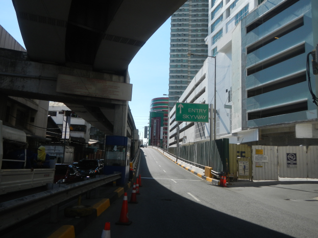 Eine Stadtstraße mit Verkehrskegeln am Rand, Fahrzeuge auf der Straße, eine Brücke links, Gebäude mit Namensschildern auf beiden Seiten, Laternenmäste und ein Himmel im Hintergrund.
