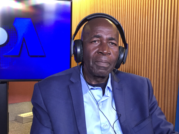 Ein Mann in Anzug und Kopfhörern sitzt vor einem Fernseher und gestikuliert, als würde er ein Gespräch führen, mit einer hölzernen Wand im Hintergrund.