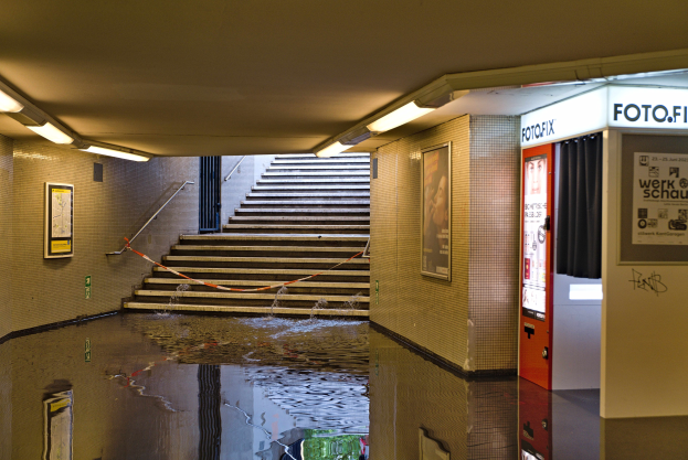 Eine überflutete U-Bahn-Station in Berlin mit Wasser auf dem Boden, Treppen mit Geländern, gerahmten Fotos an den Wänden, Deckenlampen und einem Getränkeautomaten.