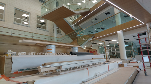 Ein großer Raum voller verschiedener Möbel- und Baumaterialien, darunter Holzplanken, Leitern, Pfeiler, Glasgeländer, Treppen, Deckenleuchten und Glasfenster auf der linken Seite, scheint renoviert zu werden.