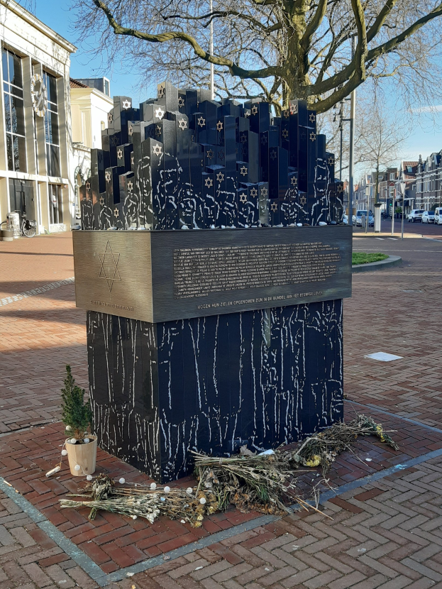Denkmal in einem Stadtplatz für Holocaust-Opfer, mit einem Baum davor, einem Topfgewächs auf dem Boden, verstreuten trockenen Blättern und Gebäuden, Fahrzeugen, Pfählen und Bäumen im Hintergrund.