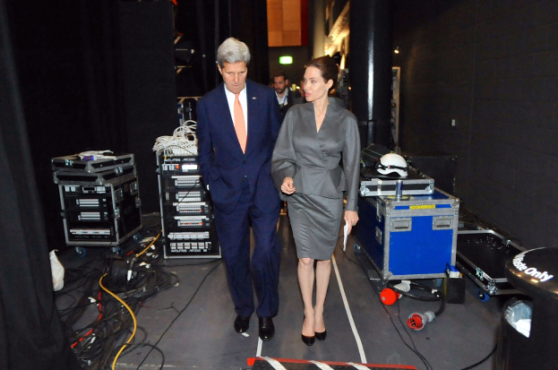 Angelina Jolie und John Kerry auf einem Boden umgeben von einer Gruppe von Menschen, verschiedenen Geräten, Kabeln, einem Helm auf einem Wagen, einem Müllcontainer, einem Vorhang, einer Säule und einer Decke mit Lampen.