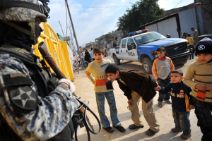 Eine Gruppe von Kindern, die vor einem Polizisten mit einer Waffe stehen, mit Fahrzeugen, Menschen, Fahrrädern und Bäumen im Hintergrund unter einem bewölktem Himmel.