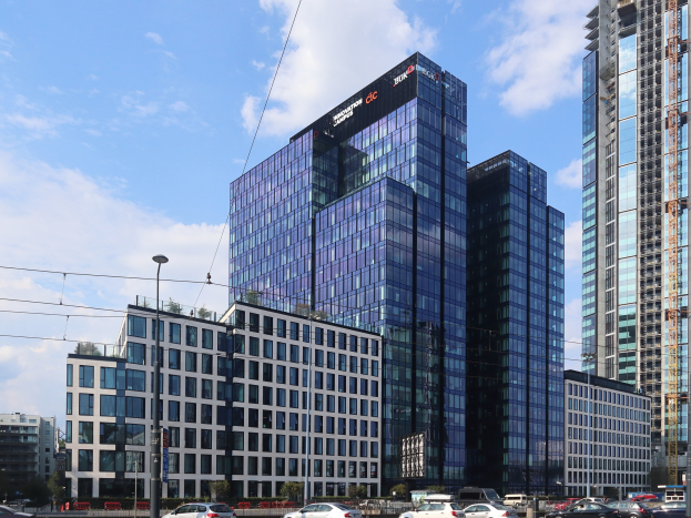 Hauptsitz der Deutschen Bank in Frankfurt, Deutschland, ein großes Gebäude mit vielen Fenstern, umgeben von Straßeninfrastruktur, Fahrzeugen, Grünflächen und einem bewölkten Himmel.