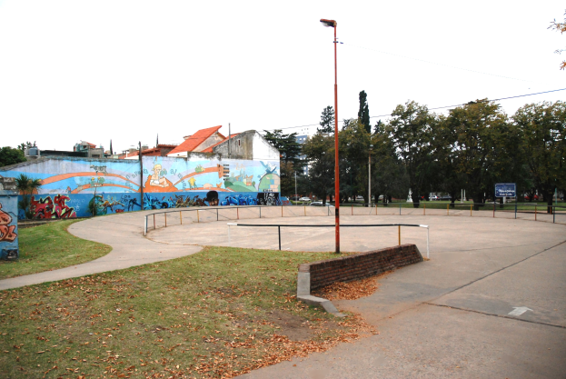 Ein Skate Park mit einer Rasenfläche, einer Wandmalerei an einem Gebäude, Laternen, Geländern und einer bemalten Wand, vor einem Hintergrund aus Bäumen, Gebäuden, Fahrzeugen und Himmel.