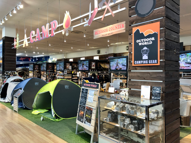 Ein Geschäft voller Campingausrüstung, darunter Zelte, Schilder, eine Glasvitrine, Fernseher, Regale mit Gegenständen, Deckenleuchten und andere Objekte.