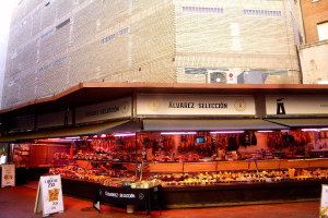 Ein belebter Markt mit verschiedenen Lebensmitteln, die unter Lichtern präsentiert werden, umgeben von Texttafeln, mit Gebäuden und einem klaren blauen Himmel im Hintergrund.