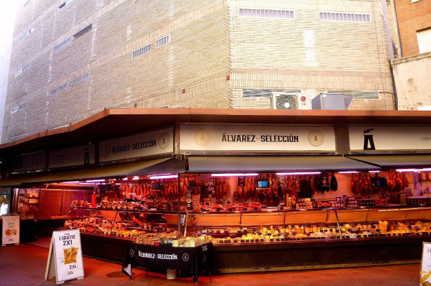 Ein belebter Markt mit verschiedenen Lebensmitteln, die unter Lichtern präsentiert werden, umgeben von Texttafeln, mit Gebäuden und einem klaren blauen Himmel im Hintergrund.