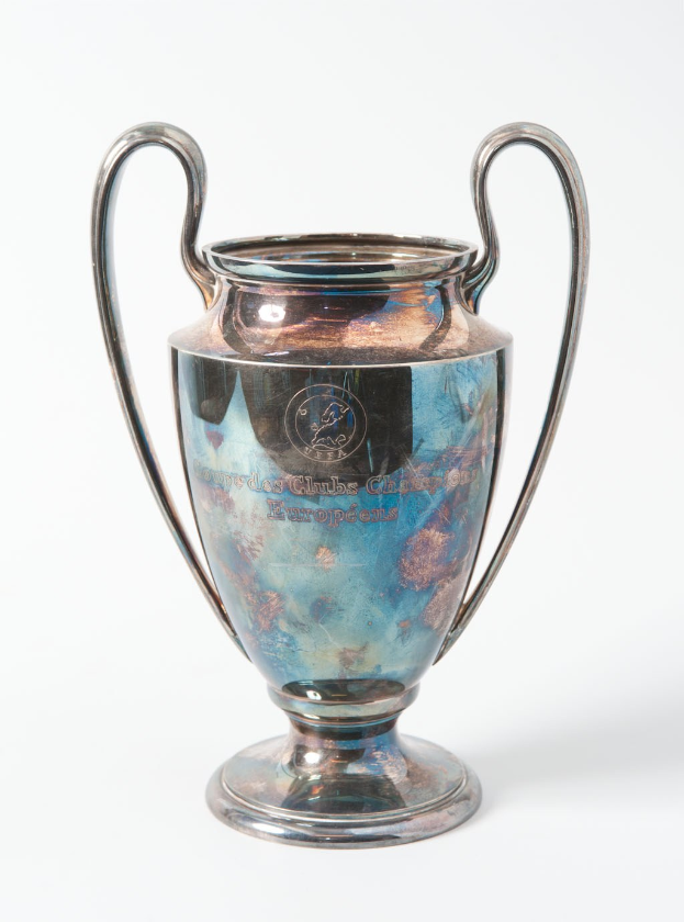 Der UEFA-Champions-League-Pokal, ein stählerner Becher mit zwei Griffen, blauen und silbernen Details und eingravierten Text, vor einem weißen Hintergrund.