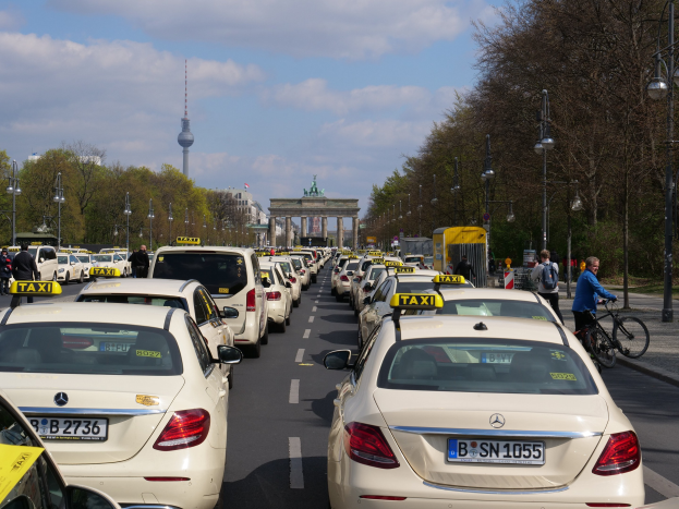 Eine lange Reihe von Taxis, die an einer belebten Straße in Berlin, Deutschland, geparkt sind, mit Fahrzeugen, Radfahrern und Fußgängern im Hintergrund, flankiert von Laternenpfählen und Bäumen und Gebäuden, einem Bogen und einem Turm in der Ferne unter einem bewölkten Himmel.