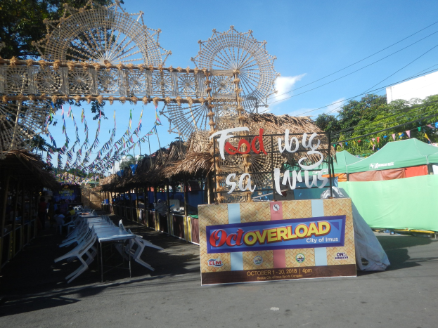 Eine Karnevalsszene mit einem großen Riesenrad in der Mitte, umgeben von Hütten, Tischen, Stühlen, einem Schild, Dekorationen, Bäumen, Überlandleitungen und einem bewölkten Himmel.