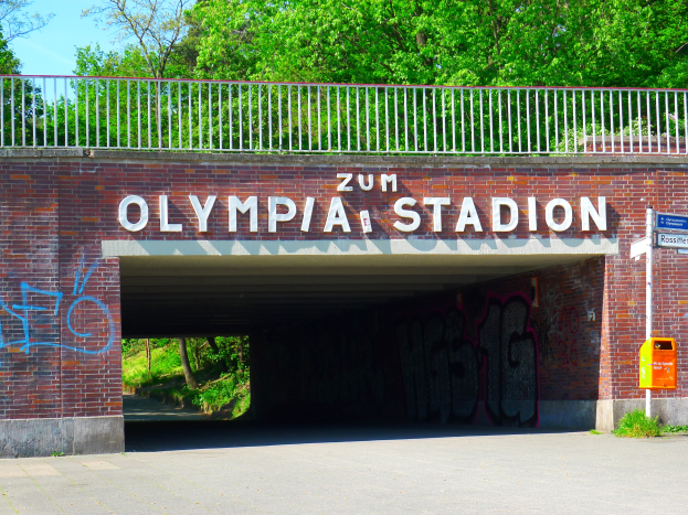 Der Eingang zum Olympiastadion in Berlin, Deutschland, mit einer Brücke mit Text, einem Metallzaun, einer Tafel, einer Box, Pflanzen, Gras, einer Gruppe von Bäumen und einem bewölkten Himmel.