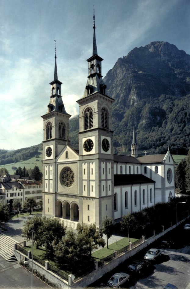 Alte Kirche mit zwei Türmen in einer Stadt, umgeben von Gebäuden, Grünflächen, Fahrzeugen und Hügeln unter einem bewölkten Himmel.