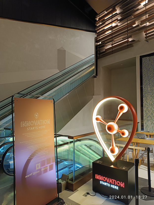 Großes Schild mit der Aufschrift "Innovation beginnt hier" in einem Gebäude mit einem Aufzug im Hintergrund, Tische und Stühle rechts und das HSTP20 Expo-Logo links.