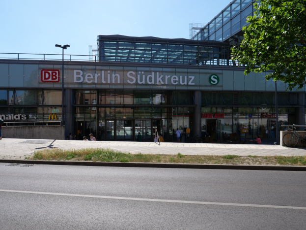Ein großes Glaswand-Bahnhof namens "Berlin Südkreuz" in Berlin, Deutschland, mit umliegender Straßeninfrastruktur, Fahrzeugen, Fußgängern, Fahrrädern, Bäumen und einem klaren Himmel.