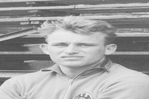 Schwarzes und weißes Foto eines Mannes in einer Jacke, der vor einem Stapel Holzplanken steht, der als legendärer deutscher Fussballer gilt.