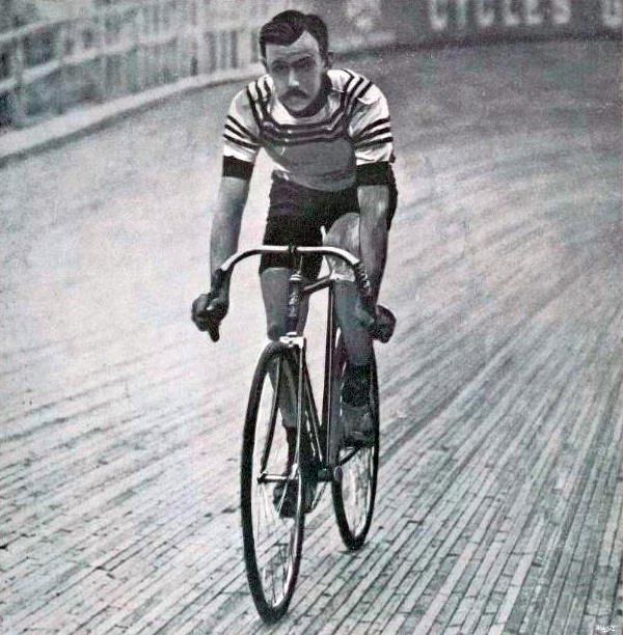 Schwarz-weiß-Foto eines Mannes, der auf einer Bahn einen Rennrad fährt, mit einem Zaun und einer Tafel im Hintergrund.
