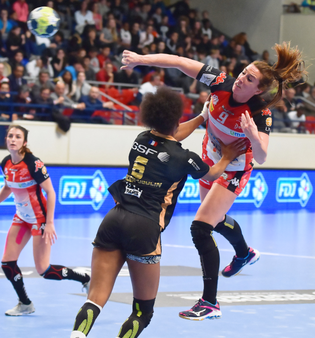 Eine Gruppe von Frauen beim Handballspielen auf einem Feld mit einem Ball in der Luft, während Zuschauer von Stühlen, Geländern und einer Wand im Hintergrund zuschauen.