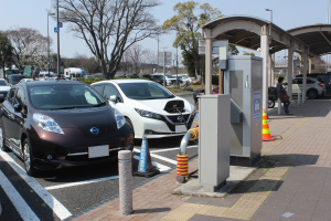 Elektroauto-Ladestation in Japan mit Autos auf der Straße, einer Person auf dem Gehweg und Infrastruktur wie Verkehrskegel, einem Schuppen, Polen, Lampen, Schildern, Bäumen und Pflanzen vor einem Himmelhintergrund.