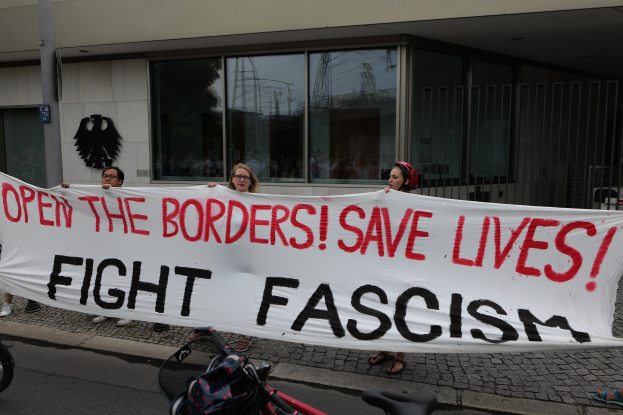 Gruppe von Menschen mit einem Transparent mit der Aufschrift "Öffnet die Grenzen, rettet Leben, kämpft gegen Faschismus" vor einem Gebäude mit Glasfenstern und einem Pfahl, mit einem Fahrrad und einer Tasche auf der Straße.