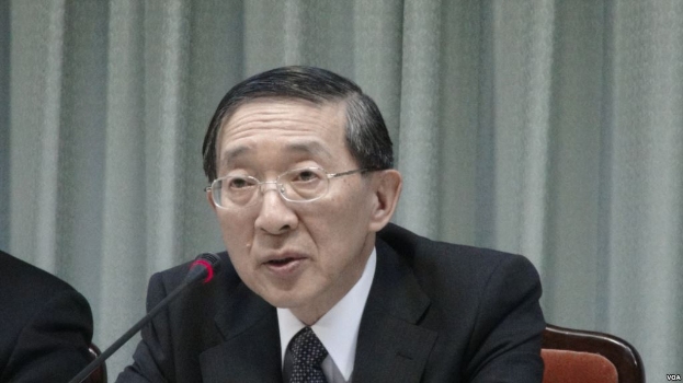 Ein Mann im Anzug und Krawatte sitzt an einem Tisch mit einem Mikrofon, trägt eine Brille und hat eine andere Person Hand auf der linken Seite und einen Vorhang im Hintergrund.