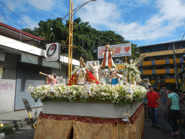 Ein Paradewagen mit Statuen, Blumen und Texttafeln, der eine von Laternenmasten, Kabeln, Gebäuden, Bäumen und einer bewölkten Himmel gesäumte Straße entlangfährt.
