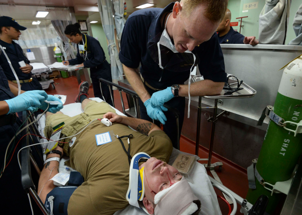 Ein Mann liegt auf einer Trage in einem Krankenhauszimmer umgeben von uniformierten und handschuhgetragenen Personen, mit einem Zylinder auf der rechten Seite und medizinischer Ausrüstung im Hintergrund, das eine U.S.-Air-Force-Covid-19-Einsatzszene zeigt.