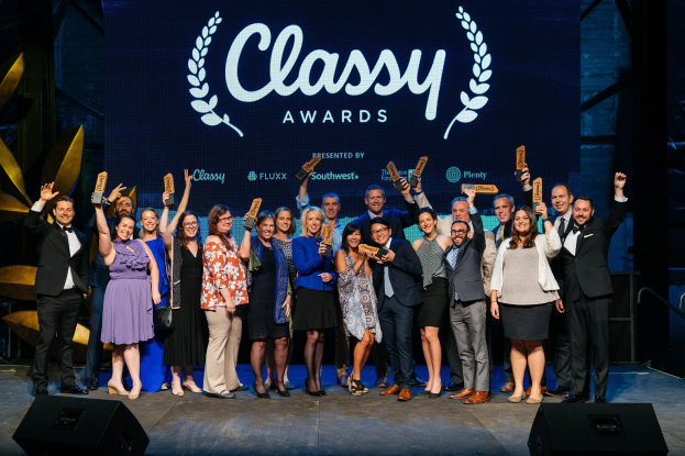 Gruppe von Menschen auf einer Bühne, die Auszeichnungen halten, mit Lautsprechern darunter und einem Bildschirm im Hintergrund, der 'Classy Awards 2019' anzeigt.