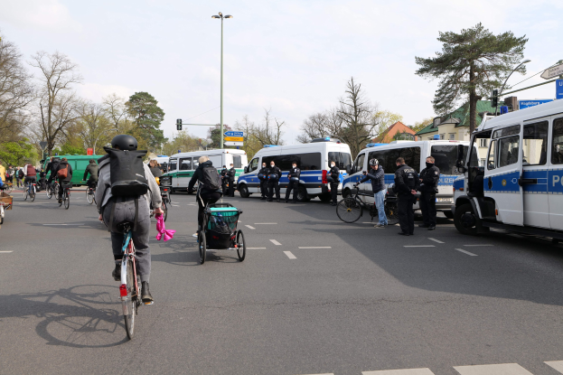 Eine Gruppe von Menschen, die auf Fahrrädern eine Straße entlangfahren, mit Polizeiwagen, einige tragen Helme und haben Taschen, andere schieben Kinderwagen, mit Bäumen, Gebäuden und Verkehrszeichen im Hintergrund.