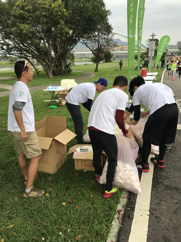 Group of people on a roadside with scattered cardboard boxes and bottles, participating in a charity run as indicated by banners, with trees, buildings, and a clear blue sky in the background.