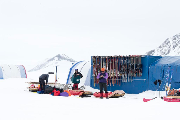 Drei Personen stehen auf einer schneebedeckten Landschaft mit verstreuten Taschen, Zelten mit Skiern dahinter und schneebedeckten Hügeln im Hintergrund bei klarem Himmel.