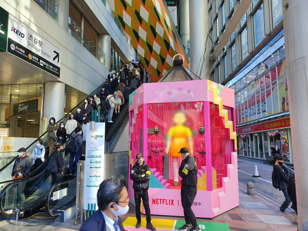 Eine Gruppe von Menschen steht vor einem Gebäude mit Glaswänden, Säulen und Texttafeln, einige tragen Masken, während hinter ihnen eine Rolltreppe mit Menschen nach oben fährt; ein pinker Kasten in der Mitte zeigt Schaufensterpuppen und trägt die Aufschrift "Netflix's neuer Netflix-Stand in Tokio."