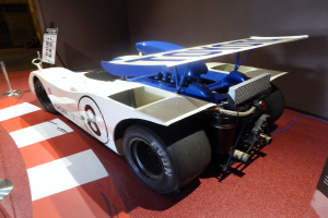 Ein weiß-blauer Rennwagen steht in einem Museum ausgestellt, umgeben von Absperrbändern und Informationsschildern, mit einer Wand im Hintergrund, die auf das Goodwood Festival of Speed hinweist.