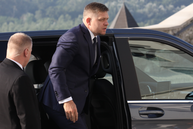 Ein Mann in einem schwarzen Anzug und Krawatte steigt aus einem Auto aus und steht neben einer anderen Person, mit Bäumen und einem Gebäude im Hintergrund.