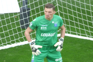 Ein Torwart in grüner Uniform steht vor einem Fußballtor auf saftigem Grün, trägt Handschuhe.