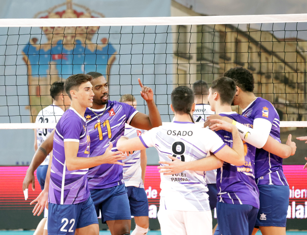 Eine Gruppe von Männern, die Volleyball auf einem Court mit einem Netz spielen, ein Banner mit Text im Hintergrund und eine Treppe mit Gelöndern.