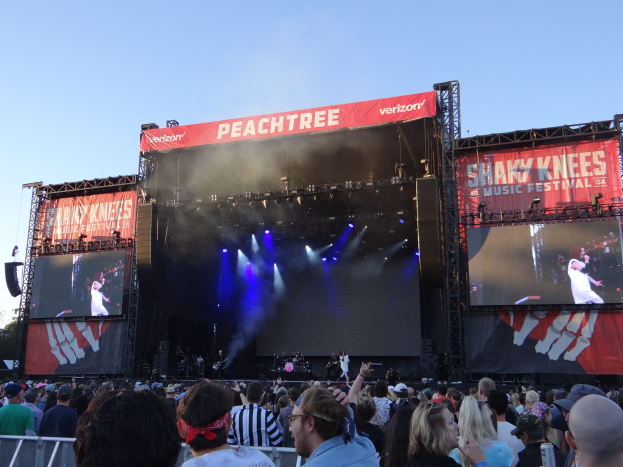 Eine große Menschenmenge steht vor einer hell beleuchteten Bühne auf dem Shaky Knees Music Festival, wo Musiker unter Bannern mit Text spielen, mit einem Geländer im Vordergrund und einem klaren blauen Himmel im Hintergrund.