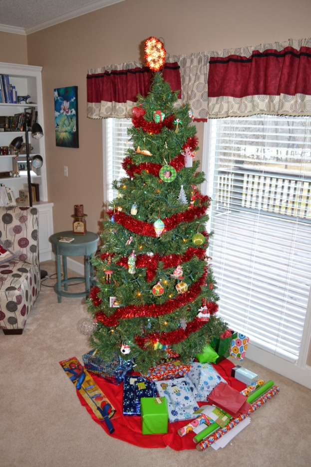Ein Weihnachtsbaum mit Geschenken davor, eine Couch, ein Tisch, ein Bücherregal, eine Lampe mit Stange und ein Bild an der Wand, alles vor einem Hintergrund von Fenstern mit Vorhängen.