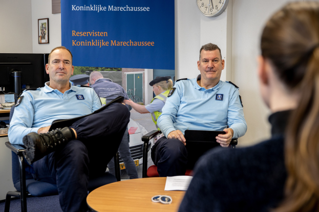 Zwei Polizeibeamte sitzen an einem Tisch mit einer Frau, umgeben von Papieren und Gegenständen, mit einem wandmontierten Banner, einer Uhr und Fotorahmen im Hintergrund und einem weiteren Tisch mit einem Monitor und Zubehör links daneben.