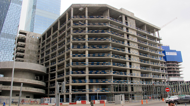 Großes Bauprojekt in einer Stadt mit Wolkenkratzern, Fahrzeugen, Fußgängern, Verkehrsinfrastruktur und Kränen unter einem klaren blauen Himmel.
