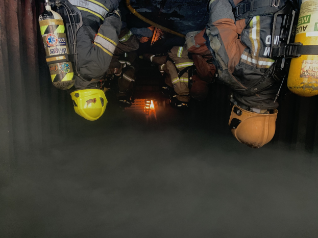 Feuerwehrleute in Schutzausrüstung und Luftzylindern arbeiten in einem raucherfüllten Tunnel mit einer Lichtquelle im Hintergrund.