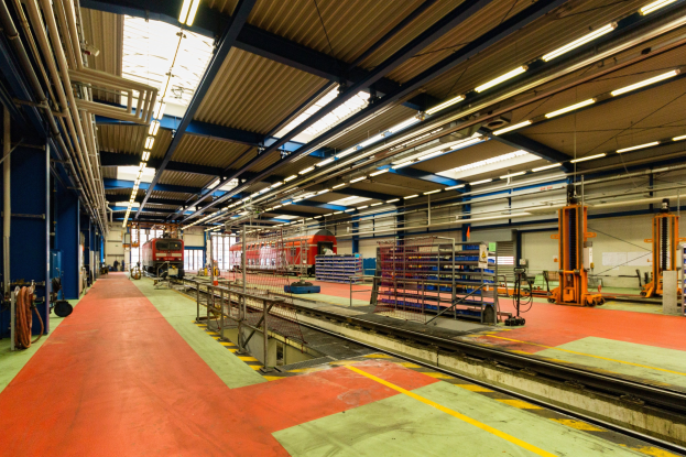 Großer Fabrikinnenraum mit rotem Teppichboden, Eisenbahnschienen, Geländern, Industriemaschinen, Polen, Lichtern und einem auf der Schiene sichtbaren Zug im Hintergrund.