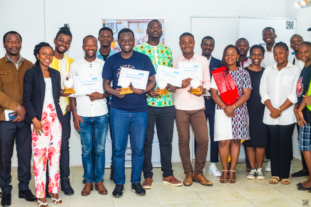Eine Gruppe strahlender Menschen hält Urkunden in der Hand und steht zusammen in einem Raum, mit einem Tisch voller Gegenstände rechts und Tafeln und einem Plakat im Hintergrund, feiernd die WIK Awards 2019.